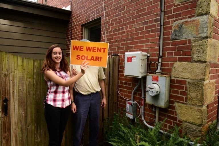 We Want Solar homeowner image from Solar United Neighbors Image of a homeowner in front of their house holding a sign "we want solar"