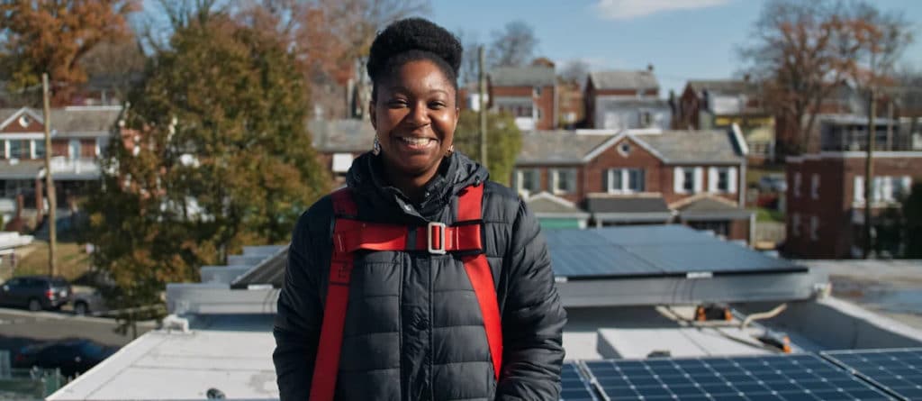 Younger African-American person standing on top of a residential building with a solar panel array installed.
